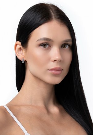Frau mit langen schwarzen Haaren, die silberne tropfenförmige Ohrringe trägt, mit dezentem Make-up und einem weißen Top mit Spaghettiträgern, leicht nach rechts gerichtet vor weißem Hintergrund.