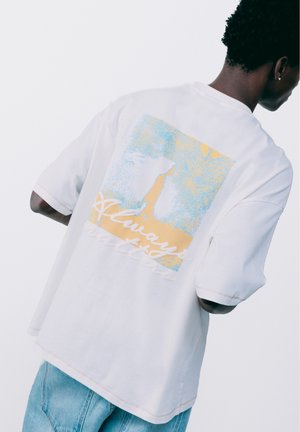 Personne portant un t-shirt blanc avec un graphisme bleu et jaune d'un chien debout et le texte "Always better" au dos, associé à un jean bleu clair.