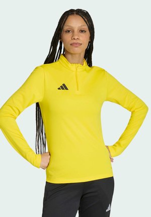 Kobieta z długimi warkoczami, ubraną w żółtą sportową bluzę z długim rękawem i czarne spodnie z logo Adidas, stojąca z rękami na biodrach.