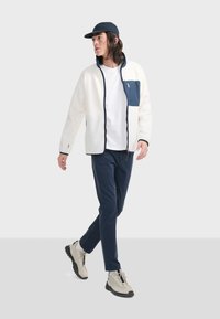 Weißer Fleece-Jacke mit marineblauen Akzenten, Reißverschlusstasche und hohem Kragen, kombiniert mit dunklen Hosen und beigen Sneakers. Modell läuft.