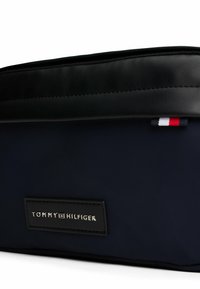 Borsa da toiletta in nylon blu navy con parte superiore in faux leather nera, forma rettangolare, etichetta con logo argentato e piccolo tag di accento rosso, bianco e blu.
