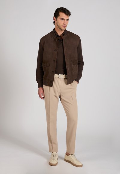 Homme portant une veste marron foncé boutonnée, une chemise foncée, un pantalon beige avec une ceinture tissée blanche et des baskets beiges, debout devant un fond uni.