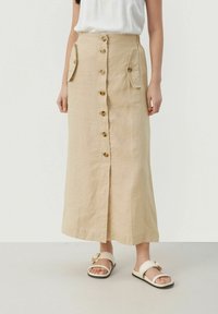 Beige linn midi kjol med framknäppning, sidofickor och rak passform. Buren med ljusa sandaler med guldförzinkring.