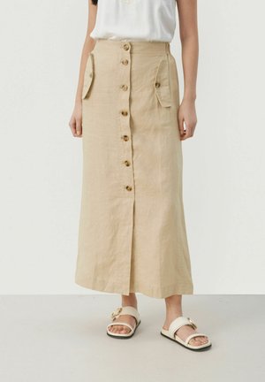 Jupe midi en lin beige avec fermeture à boutons sur le devant, poches latérales et coupe droite. Portée avec des sandales légères dotées d'un accent doré.