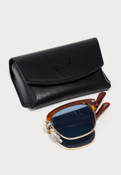 Ochelari de soare pătrați maro și aurii pe un etui din piele neagră cu logo embosat. Ochelarii dispun de lentile închise la culoare și detalii distinctive.