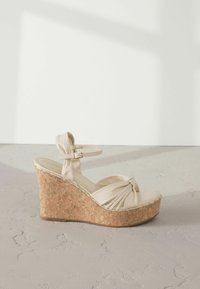 Sandalo con zeppa in pelle beige e plateau in sughero. Presenta un design frontale annodato e una cinghia regolabile alla caviglia. Texture liscia e punta arrotondata.