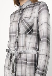 Femme portant une robe-chemise à manches longues à carreaux noirs et blancs avec des boutons, une poche poitrine, un col et une ceinture à nouer à la taille.