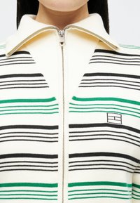 Pulover cu fermoar tricotat, de culoare crem, cu dungi verzi și negre. Prezintă un guler lat și un mic logo pe pieptul stâng. Material texturat.