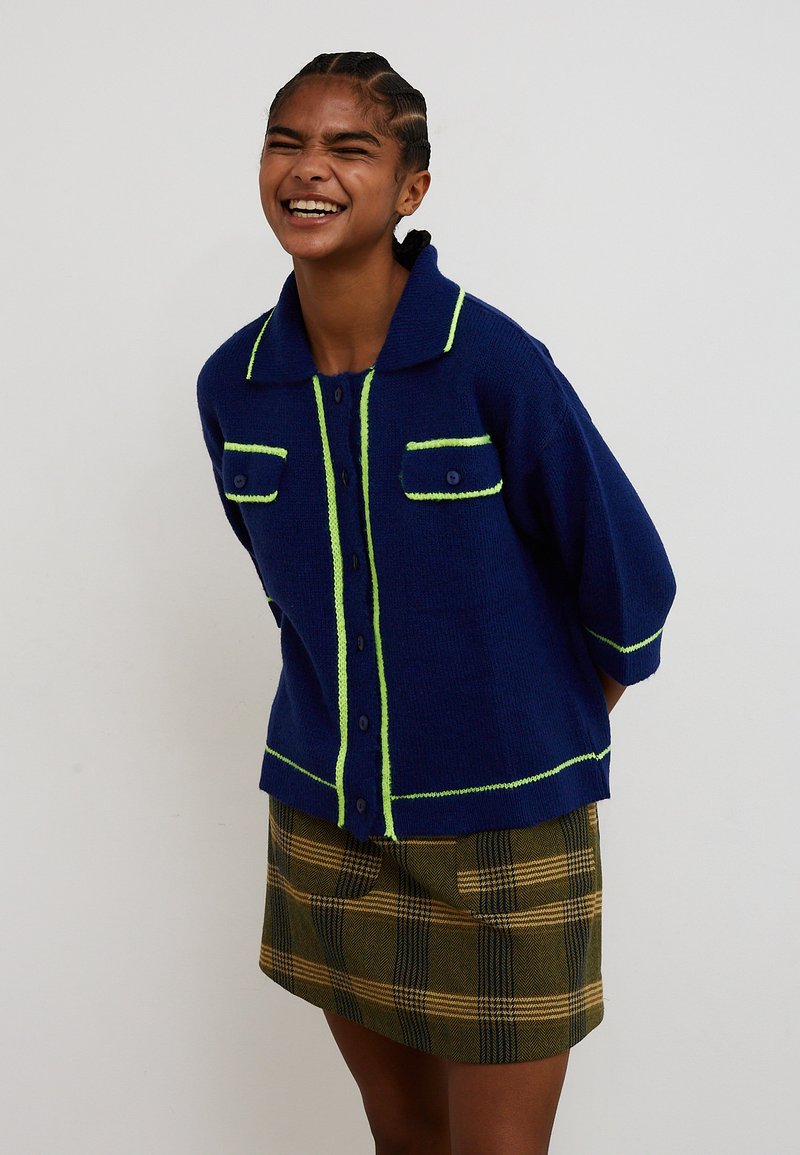 Persona sorridente con capelli intrecciati indossa un cardigan blu con bottoni e bordo lime e una gonna a quadri verde su uno sfondo semplice.