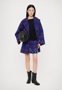 Mulher vestindo um casaco e saia xadrez em azul e vermelho com detalhes em pérola, suéter preto, botas pretas, segurando uma grande bolsa preta.