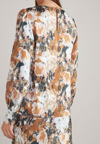 Femme portant un chemisier à manches longues avec un imprimé floral abstrait en marron, blanc et noir, et un détail en forme de goutte d'eau au niveau de l'encolure arrière.