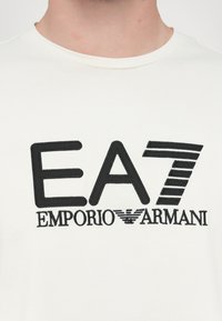 Hvit bomull t-skjorte med svart brodert logo "EA7 EMPORIO ARMANI" i fete bokstaver og et stripet design på "7."