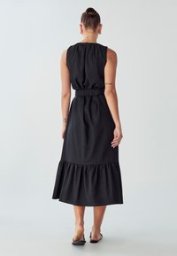 Donna con tatuaggi che indossa un vestito midi nero senza maniche con orlo arricciato e sandali neri a slip, in piedi davanti a uno sfondo semplice.