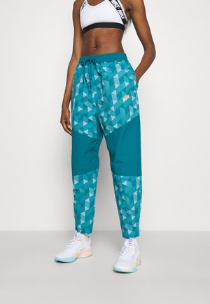 Person iført teal Nike joggings med geometrisk mønster, hvid Nike sports-bh og hvide sneakers, stående mod en ensfarvet baggrund.