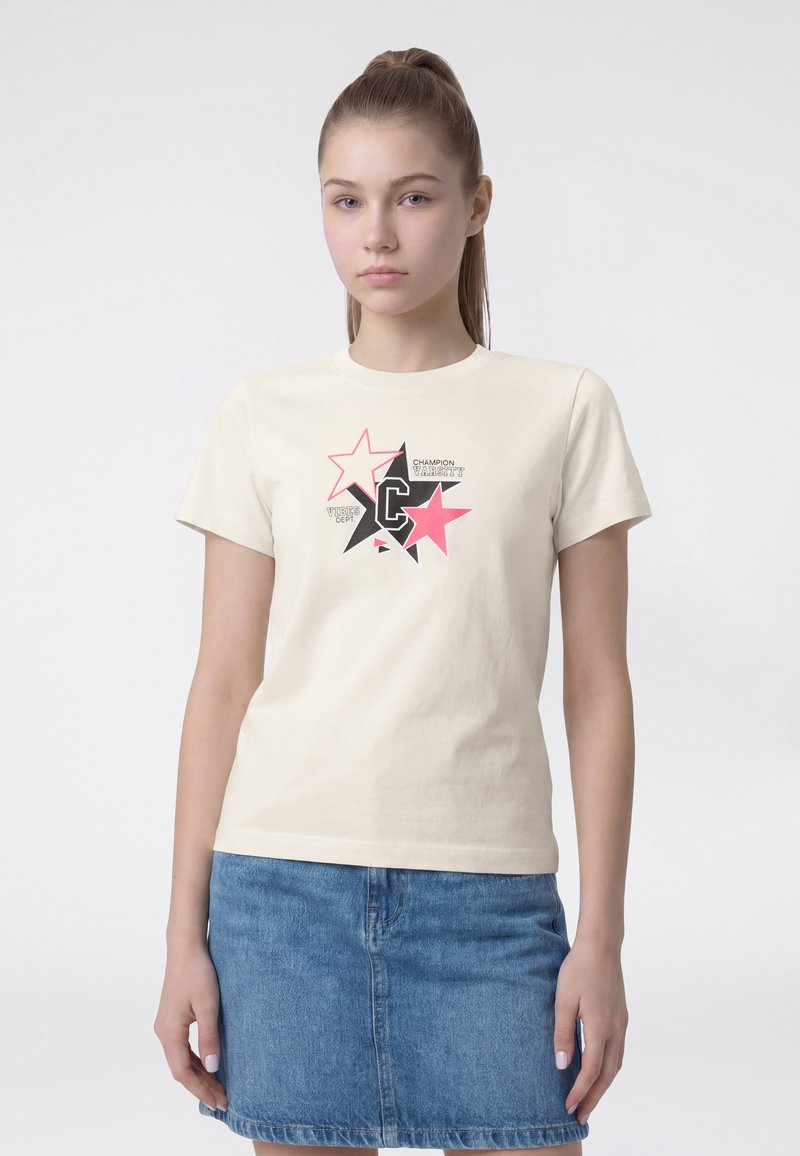 Νεαρή γυναίκα με αλογοουρά φοράει κρεμ t-shirt με γραφικό αστέρι και τζιν φούστα, στέκεται μπροστά σε απλό φωτεινό φόντο.