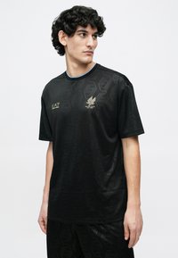 Melnas sporta krekls ar sešstūra rakstu, apaļu kaklu un mazu zelta logotipu kreisajā krūtīs. Īsas piedurknes, gluda tekstūra.