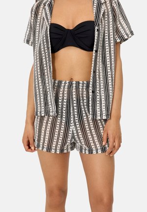 Mujer vistiendo un top bandeau negro con una camisa abierta a juego con estampado y shorts de cintura alta en rayas negras, blancas y beige.