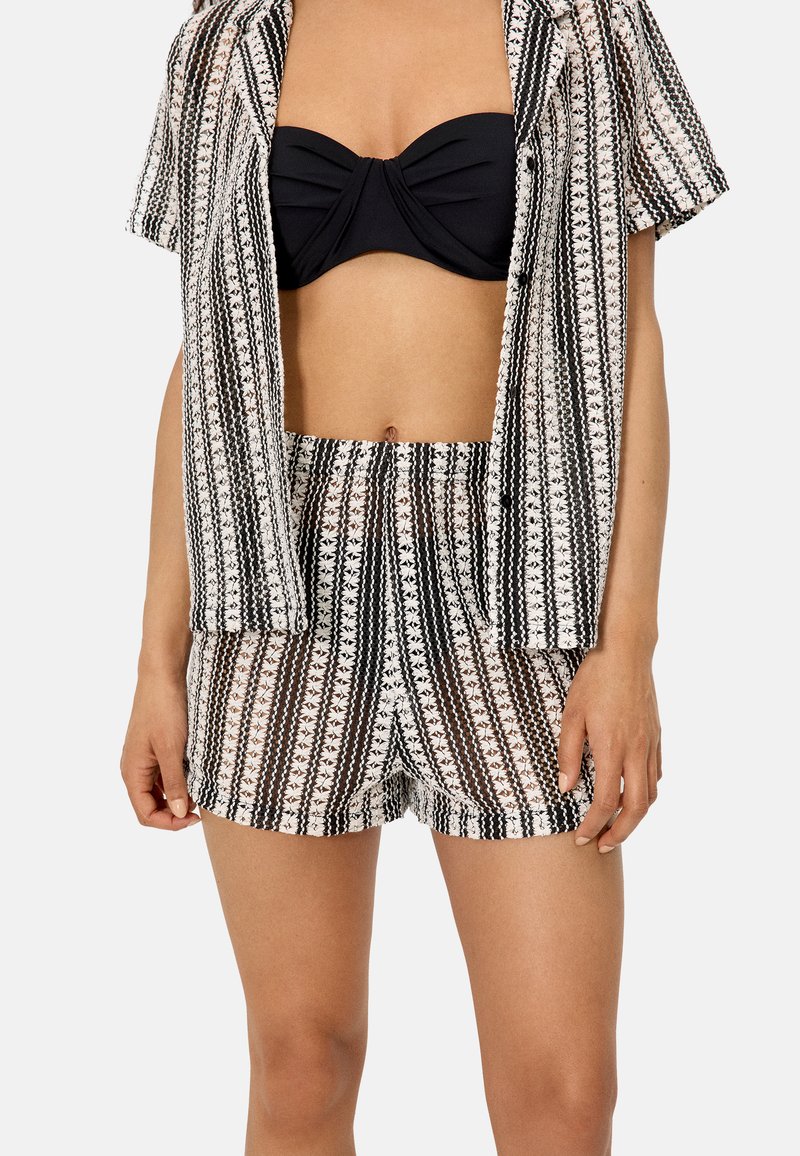 Mujer vistiendo un top bandeau negro con una camisa abierta a juego con estampado y shorts de cintura alta en rayas negras, blancas y beige.