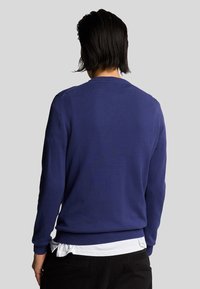 Felpa blu navy con maniche lunghe, scollo rotondo, polsini e orlo a costine, texture morbida, indossata sopra una camicia bianca per un look a strati.