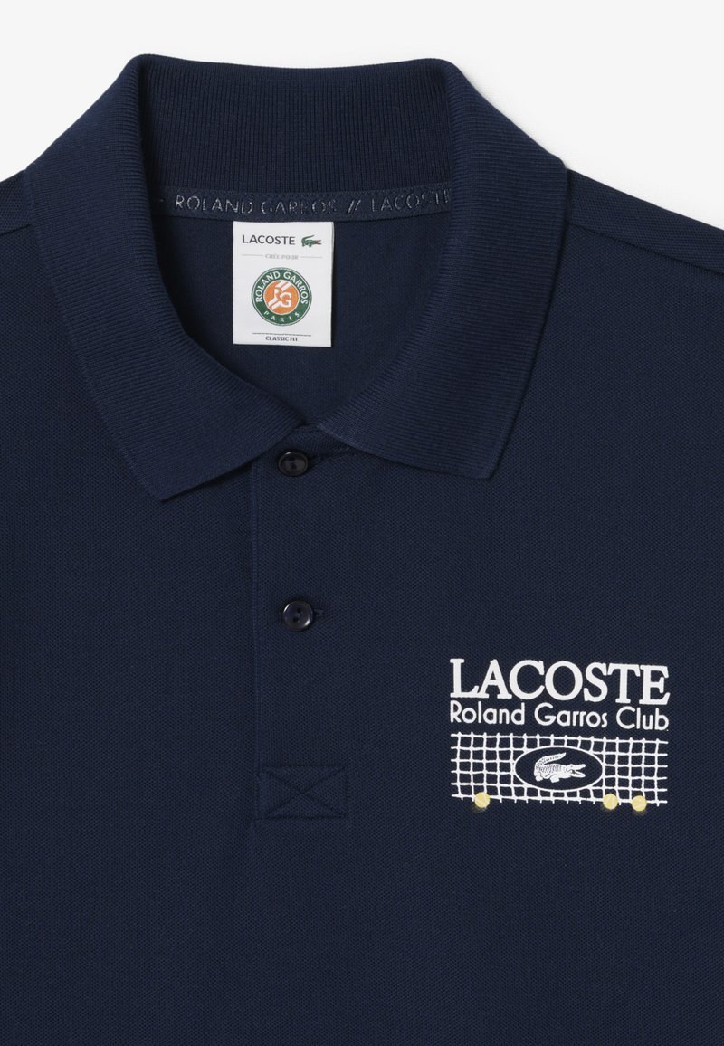 Polo bleu marine avec les logos Lacoste et Roland Garros Club, comprenant des boutons et un motif brodé de filet de tennis avec balles sur la poitrine.