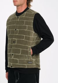 Chaleco de forro polar verde oliva con patrón de ladrillo, botones a presión y bolsillos laterales. Llevado sobre una camisa negra de manga larga.