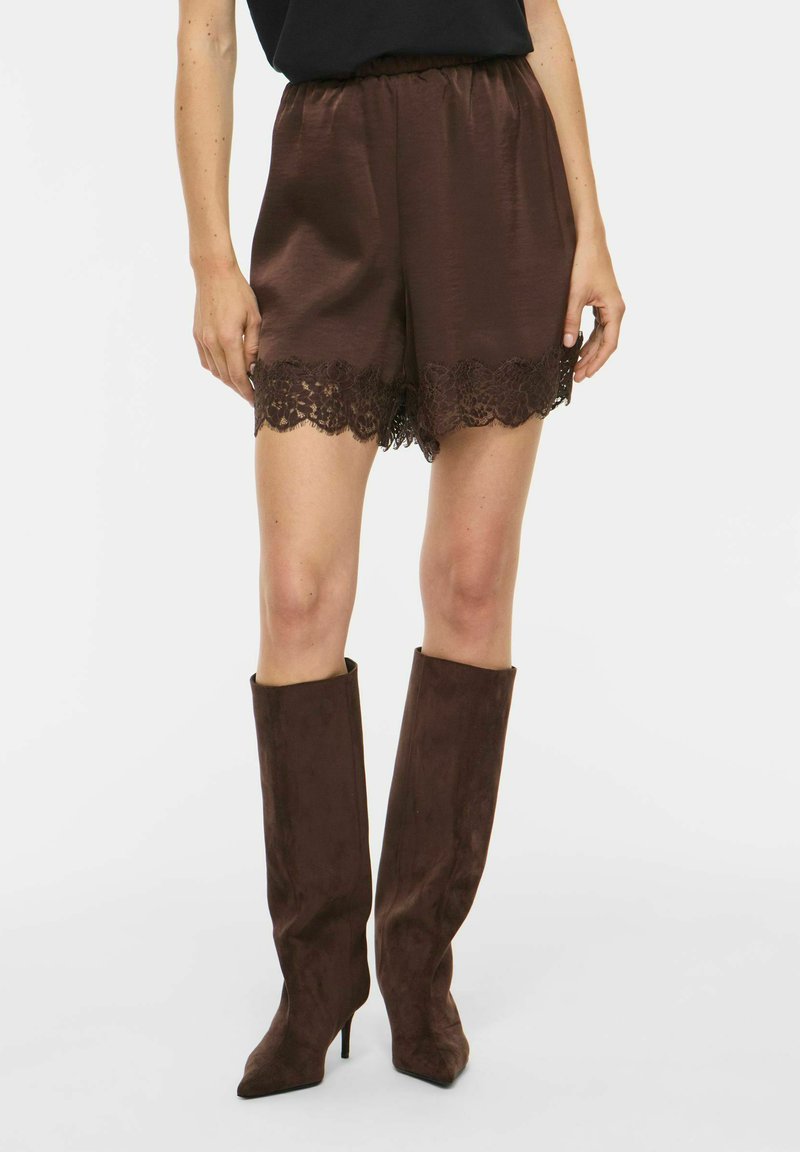 Femme portant un short en satin marron avec bordure en dentelle et des bottes hautes en daim marron foncé, debout devant un fond clair uni.