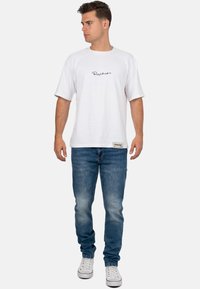Witte t-shirt met korte mouwen en zwart scriptlogo, gecombineerd met blauwe denim jeans en witte sneakers met rode accenten.