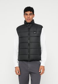 Melna pūkaina veste ar augstu apkakli, priekšējo rāv�ējslēdzēju un sānu kabatām. Gluda faktūra, quiltēts dizains ar taisnstūrveida segmentiem, logo uz krūtīm.