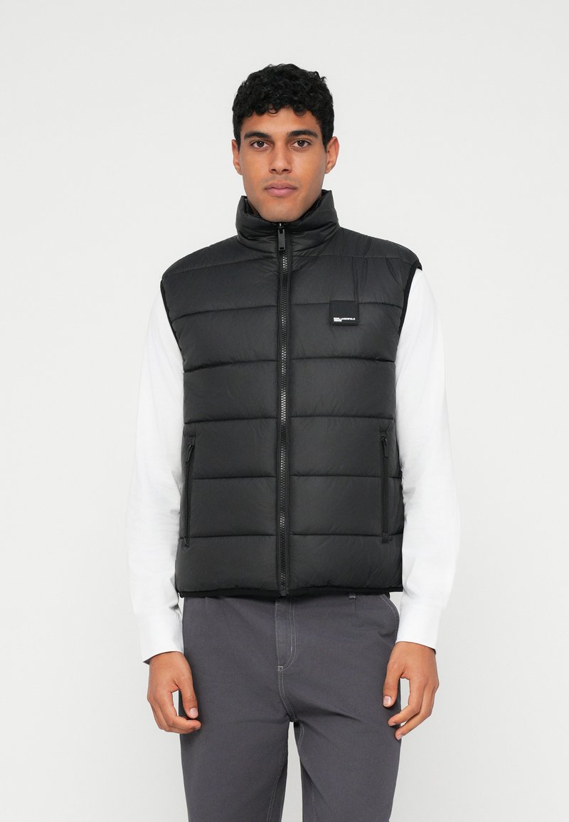 Melna pūkaina veste ar augstu apkakli, priekšējo rāvējslēdzēju un sānu kabatām. Gluda faktūra, quiltēts dizains ar taisnstūrveida segmentiem, logo uz krūtīm.