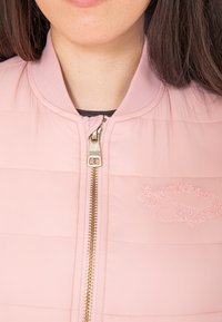 Różowa kurtka bomberka z pikowanym wzorem, z złotym zamkiem błyskawicznym i wyhaftowanym logo po lewej stronie. Ściągacz na kołnierzu.