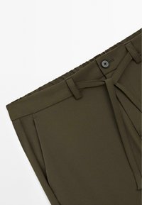 Gros plan sur la taille de pantalons vert olive avec un bouton noir, des passants de ceinture et une ceinture en tissu assorti sur fond blanc.