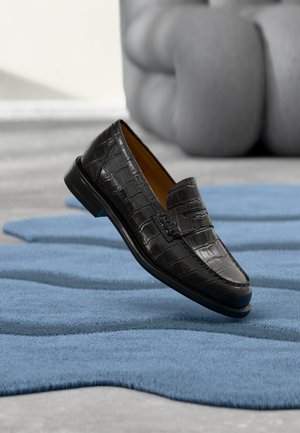 Mocassin en cuir noir à texture crocodile flottant au-dessus d'un tapis bleu de forme abstraite sur un sol gris clair.