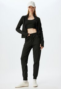 Zwart sportoutfit bestaande uit een zip-up jack en een aansluitende crop top met bijpassende joggers. Materiaal lijkt glad; geaccentueerd met een lichte pet.