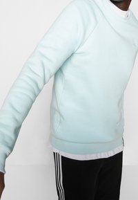 Ljust blå crewneck sweatshirt med raglanärmar, mjuk textur och en diskret logotyp på bröstet, bärs över en vit skjorta och svarta byxor.