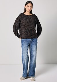 Maglietta nera a maniche lunghe con un design a stampa leopardata, abbinata a jeans in denim azzurro chiaro e sneakers beige, su uno sfondo neutro.