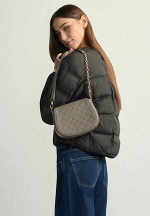 Kobieta o długich brązowych włosach ubrana w czarną puchową kurtkę i niebieskie dżinsy, trzyma na ramieniu szarą pikowaną torebkę z złotym łańcuszkiem.