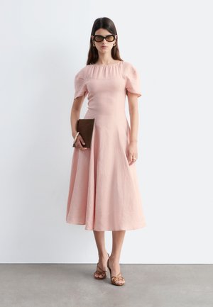 Femme portant une robe midi rose clair à manches ballon, tenant une pochette marron, portant des lunettes de soleil foncées et des talons marron à brides.