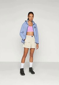 Mujer de pie vestida con una chaqueta de cremallera azul clara, top corto lila, pantalones cortos beige con cinturón, calcetines blancos y botas de senderismo negras frente a un fondo liso.