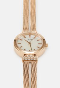 Zlati kronometr s okroglo, kovinsko ohišjem, belim številčnico in zlatimi kazalci. Mrežasta zapestnica ima okrasno zaponko z dodatnimi kristali.