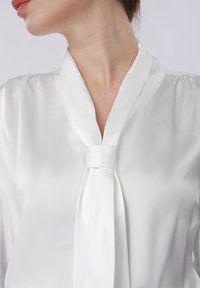 Femme portant un chemisier en soie blanc avec un large détail de cravate, vue des épaules au menton, tête tournée vers la gauche.