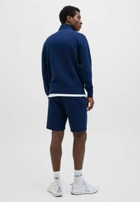 Sudadera con cremallera de color azul marino y cuello alto, combinada con pantalones cortos a juego. Presenta un diseño minimalista y una textura suave.