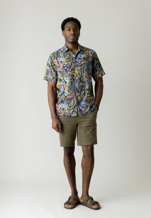 Mand iført en farverig kortærmet skjorte med blomster- og dyreprint, olivengrønne shorts og brune sandaler, stående foran en ensfarvet baggrund.