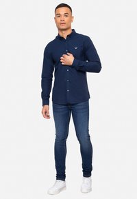 Camisa azul marina de botones con mangas largas, diseño ajustado y un pequeño logo en el pecho, combinada con jeans ajustados oscuros y zapatillas blancas.