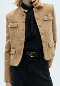 Veste courte texturée beige avec poches à rabat boutonnées sur un chemisier noir à col avec nœud, associée à un pantalon noir taille haute avec ceinture.