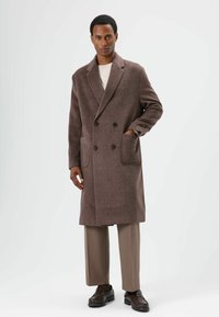Manteau croisé en mélange de laine marron avec de grandes poches plaquées et de larges revers, associé à un pantalon beige et des chaussures foncées.