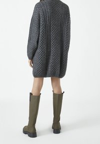 Pull en tricot gris surdimensionné avec un motif texturé et des manches longues, associé à des bottes en caoutchouc vert olive montant jusqu'aux genoux avec des accents noirs.