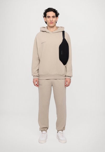 Beżowy bawełniany hoodie z kieszenią kangurzą, w połączeniu z dopasowanymi dresami. Czarna torba na ramię spoczywa na lewym ramieniu. Białe sneakersy.