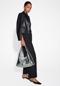 Giubbotto corto in pelle nera con dettagli silver, abbinato a un completo nero. Una grande borsa metallica silver è tenuta in una mano.