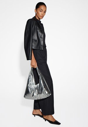 Schwarze Lederkurzjacke mit silbernen Details, kombiniert mit einem schwarzen Outfit. Eine große metallisch-silberne Tasche wird in einer Hand gehalten.