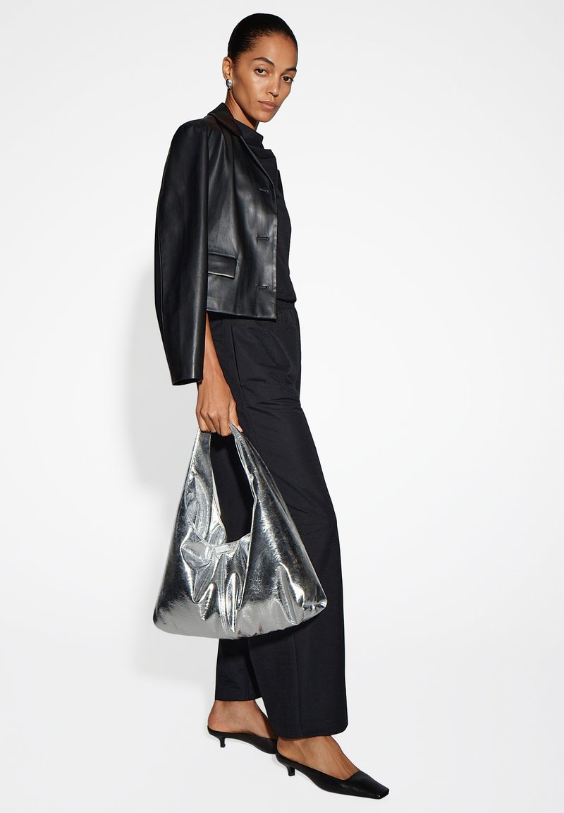 Giubbotto corto in pelle nera con dettagli silver, abbinato a un completo nero. Una grande borsa metallica silver è tenuta in una mano.